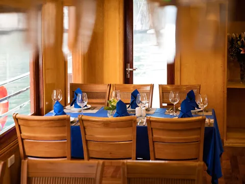 A wood-paneled boat dining room with a table set in blue cloth and napkins, wine glasses and wooden chairs, with a life ring outside the window and a flower vase on a shelf at right.