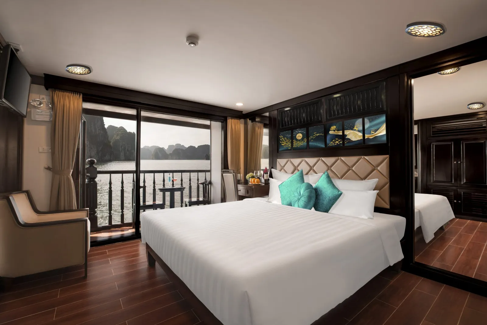 A cruise cabin with a large white bed, teal cushions, dark wood panelling and a mirrored wardrobe, with glass doors opening onto a balcony that looks out over water and limestone karst islands.
