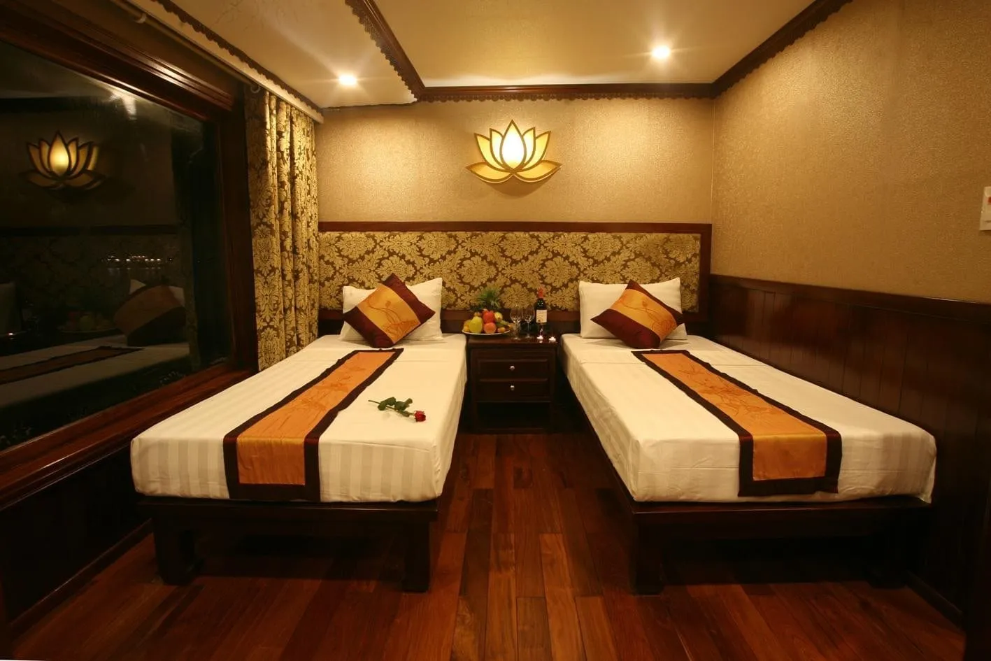 A cabin with two twin beds with orange and brown runners, a bedside table with a fruit plate and wine, a lotus-shaped wall lamp, dark wood floor and paneling, and a large window on the left.