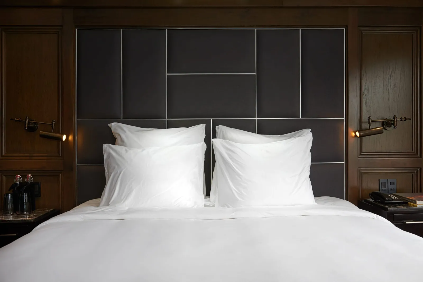 A made bed with white linens and four white pillows against a dark padded panel headboard, flanked by dark wood paneling, brass reading lights and bedside tables with a phone and glasses.