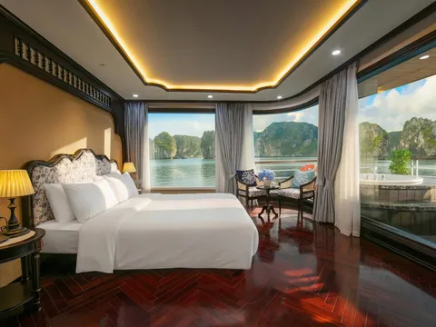 A cruise cabin with a white double bed, dark herringbone wood floor, and floor-to-ceiling windows overlooking calm water and limestone karst islands, with a private jacuzzi on the balcony.