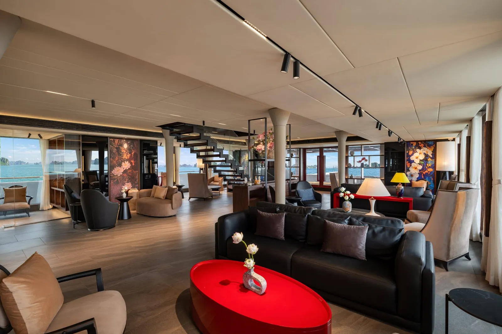 A spacious cruise-ship lounge with black leather sofas, a red oval coffee table, floral panels, a floating staircase, and large windows showing calm water and distant karst islands.