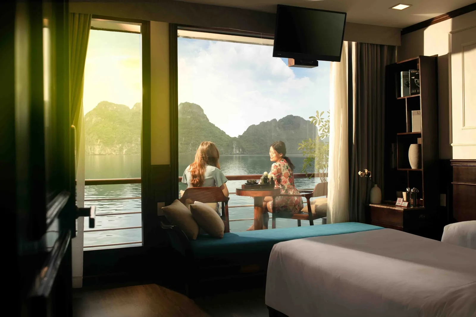 Interior of a cruise cabin with a bed and shelves, looking through open glass doors to a balcony where two women sit at a small table facing a bay with green limestone karst peaks.