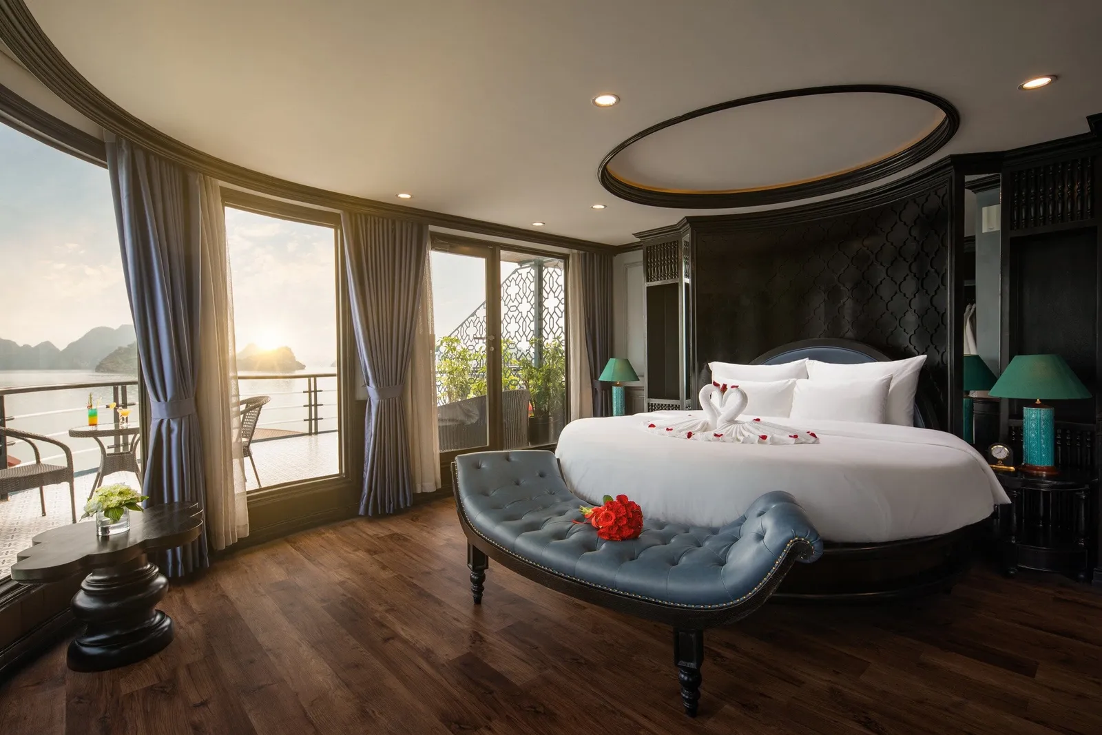A cabin with a round bed with a towel swan and rose petals, a blue tufted bench with red roses, and floor-to-ceiling windows opening to a balcony overlooking a bay with limestone karsts at sunset.