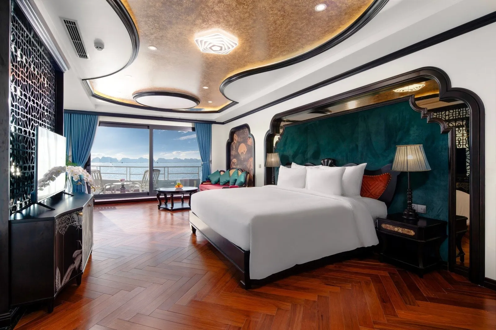 A luxurious cruise cabin with a large white bed, teal velvet headboard wall, herringbone wood floor and a sliding glass door opening to a balcony overlooking a bay with limestone karsts.