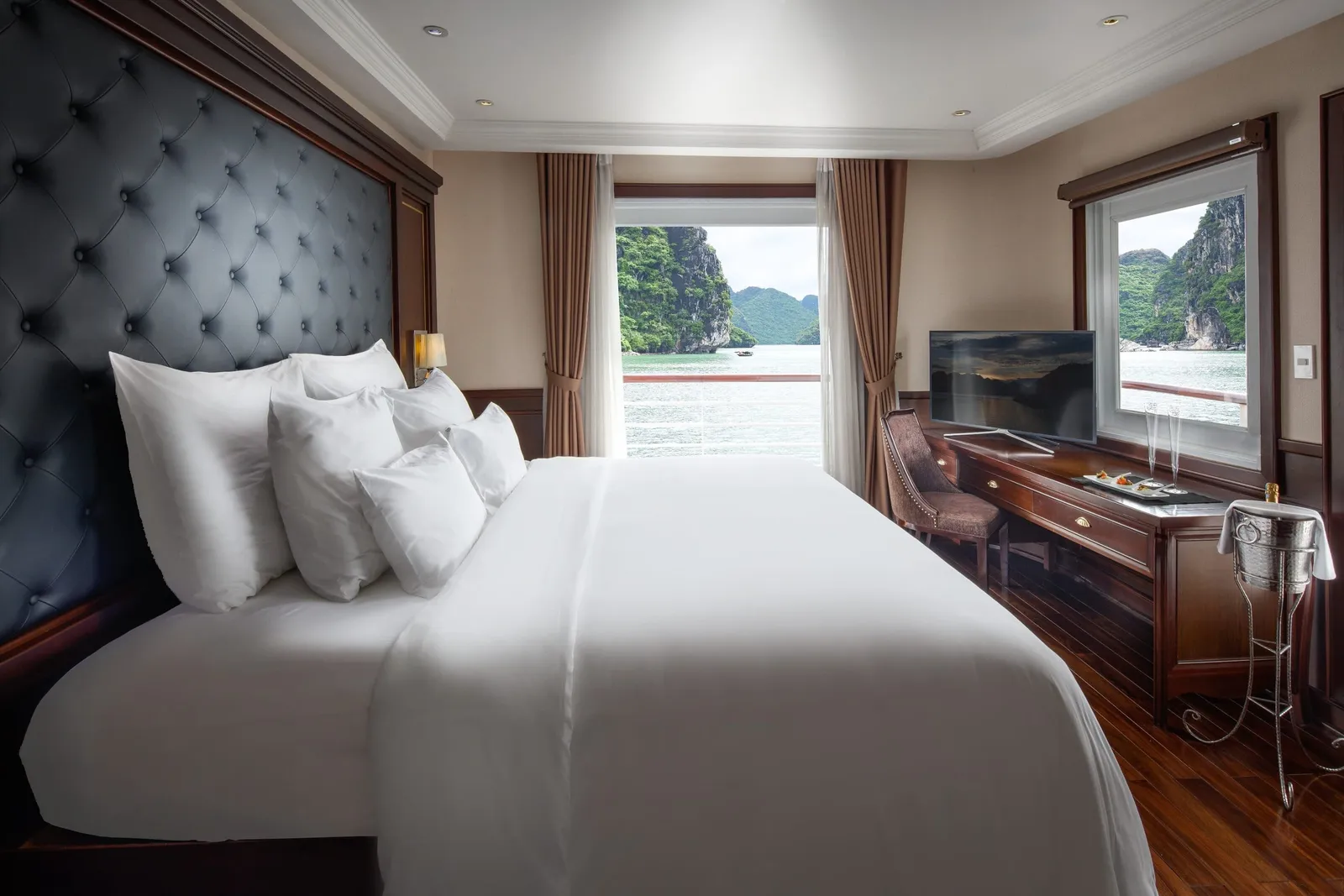 A cruise cabin with a large white bed, tufted dark headboard, wooden desk with TV and champagne bucket, and windows and open curtains showing green limestone karsts rising from the bay.