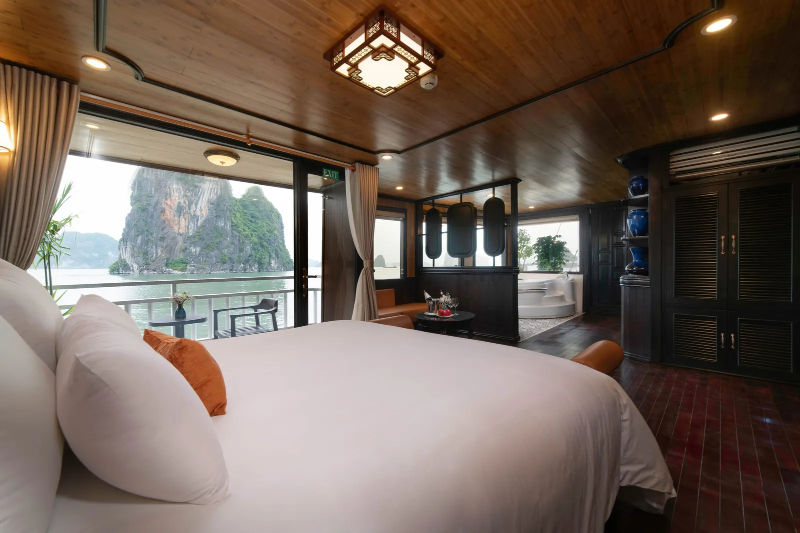 A wood-paneled cruise cabin with a large white bed, a balcony with a table and chair, a corner jacuzzi, and a limestone karst island visible across the water through the open glass doors.