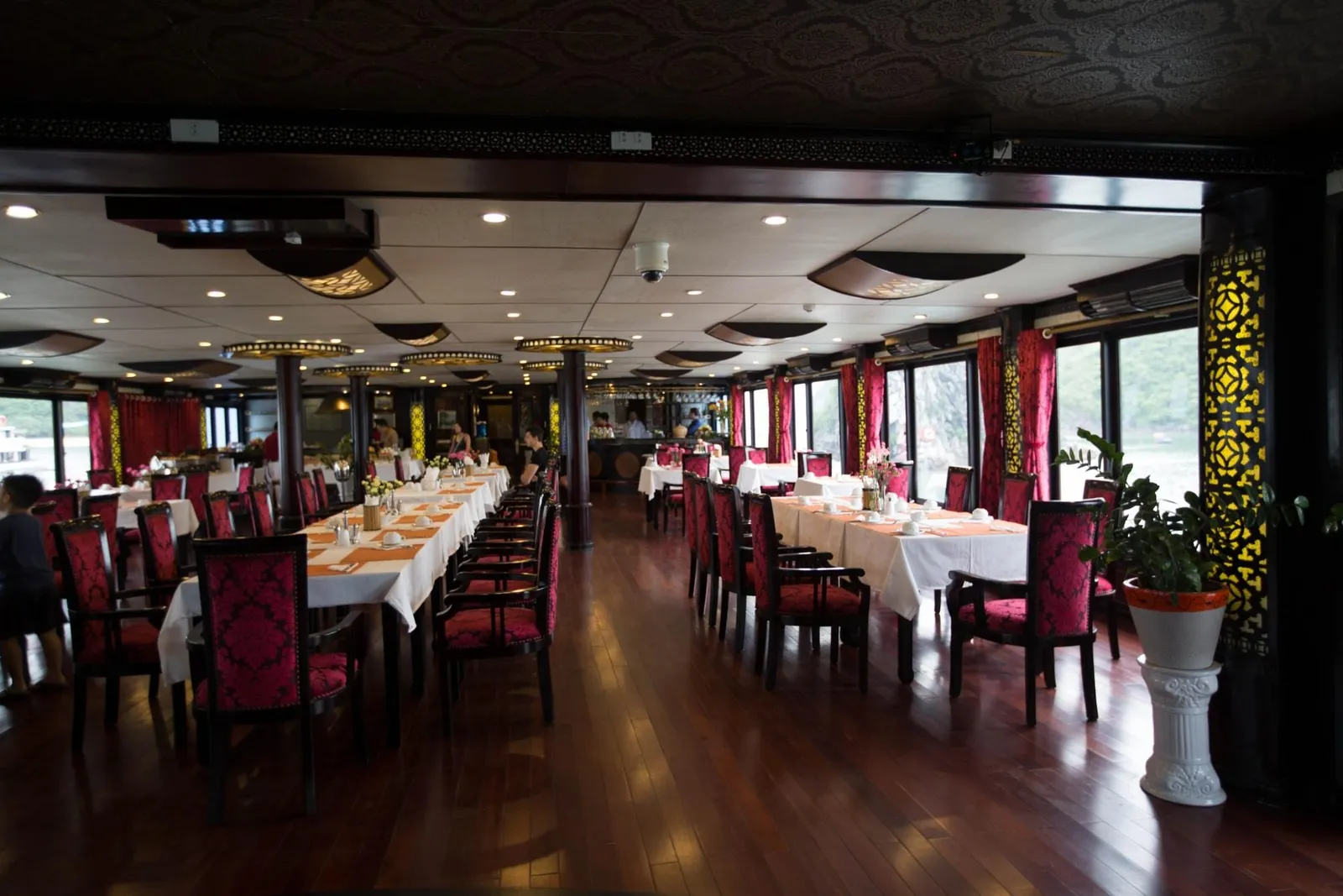 Interior dining room of a boat with long white-clothed tables, red patterned chairs, dark wood floor, a potted plant, and windows showing green karst hills and water outside.
