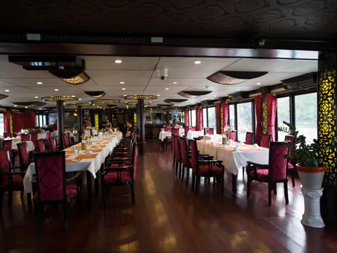 Interior dining room of a boat with long white-clothed tables, red patterned chairs, dark wood floor, a potted plant, and windows showing green karst hills and water outside.