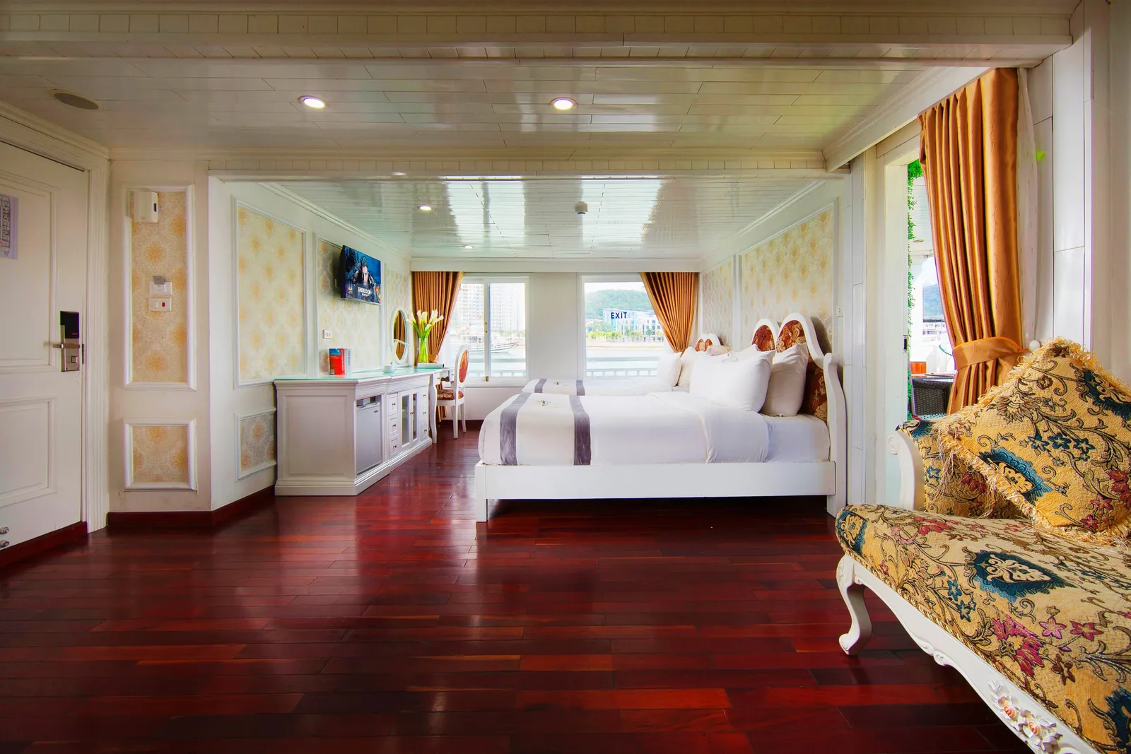 A spacious cabin with polished dark red wood floor, two white beds, ornate floral chaise lounge, gold curtains, a wall-mounted TV and windows showing the water and a green hill outside.