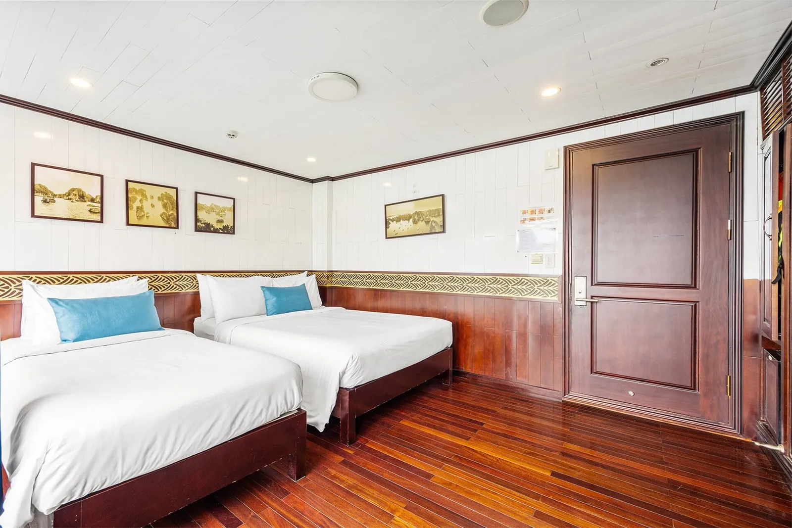 A cabin with two twin beds with white linens and blue cushions, dark wood floor and door, white paneled walls, and framed sepia prints of limestone karst bay scenery.