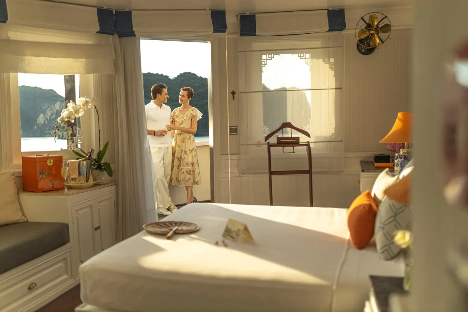 A bright cabin with a white bed, orange cushions and a window seat, with a couple holding drinks on a balcony doorway beside limestone karst hills across the water.
