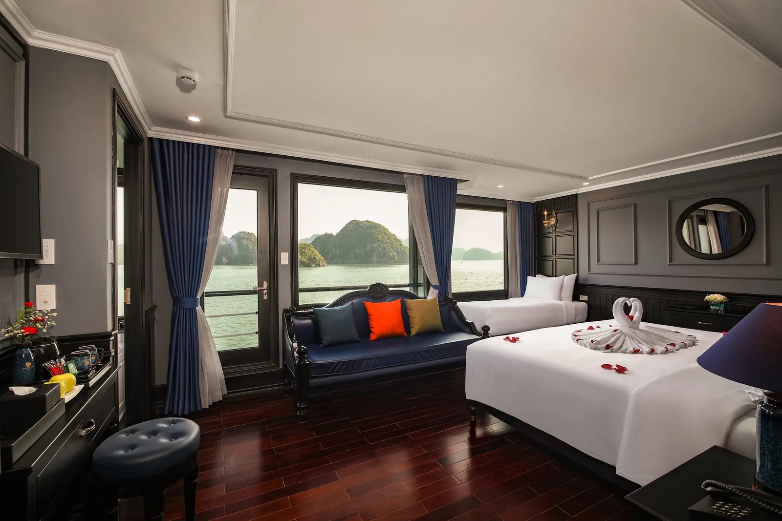 A cruise cabin with two beds, a towel-folded swan on one, a blue sofa with colorful cushions, and windows and a glass door showing green water and limestone karst islands.