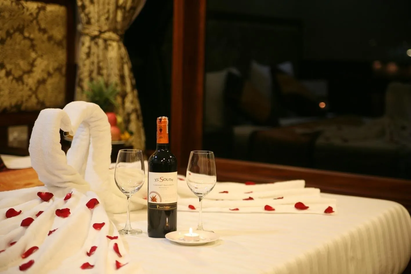 A bed decorated with a towel swan, red rose petals, a bottle of 35 South wine, two empty glasses and a lit candle, in a dimly lit cabin with gold curtains and a fruit bowl behind.