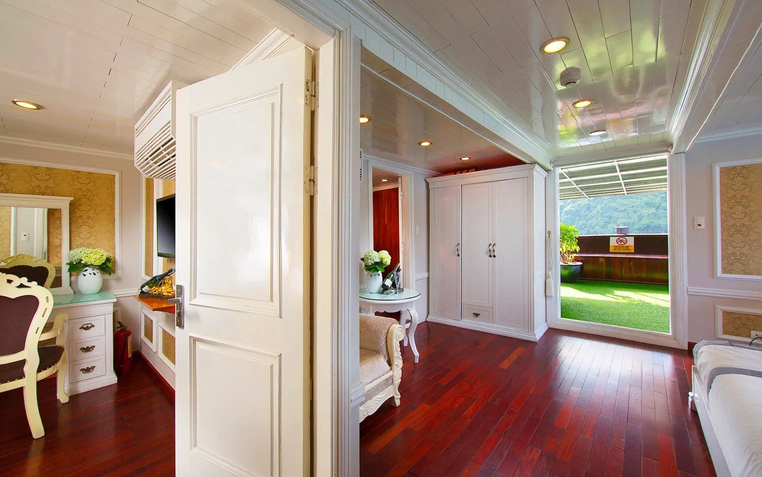 A white-paneled suite interior with dark red hardwood floors, an open white door, a vanity with flowers, a wardrobe, and an open doorway leading to a private artificial-grass terrace with a forested limestone hill behind it.