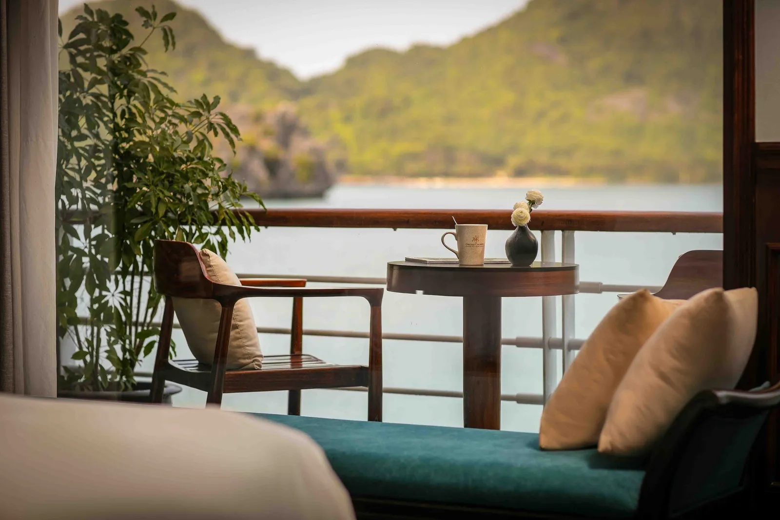 A cabin balcony with a wooden chair, round table with a logo-marked mug and a vase of white flowers, a teal daybed with cream cushions, and a potted plant, overlooking calm water and green forested karst hills.