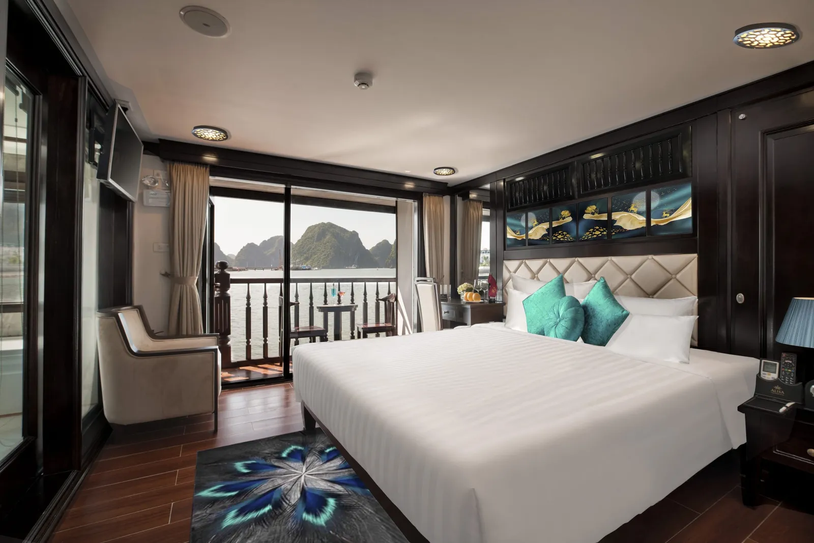 A dark-wood cruise cabin with a white double bed, teal cushions, an armchair and a balcony door opening onto a bay with limestone karst islands; a small branded logo sits on the bedside phone.