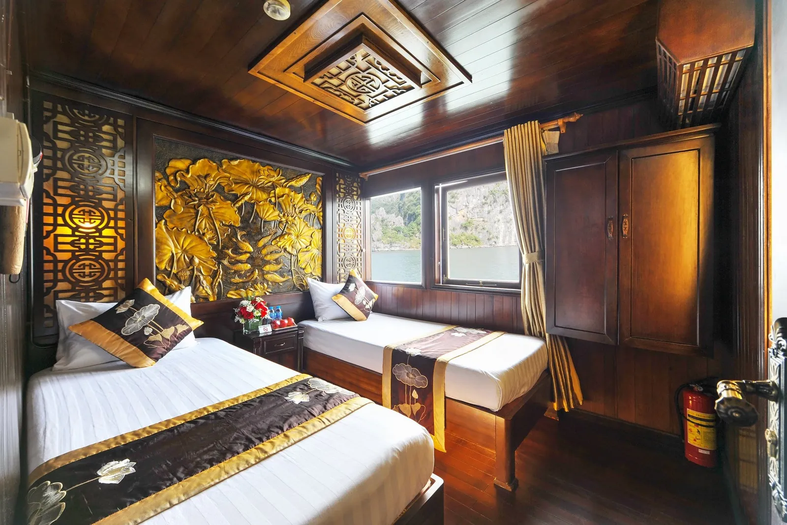 A wood-paneled twin cabin with two white beds, lotus-patterned runners, a gold carved lotus panel, a wardrobe and a fire extinguisher, with a window showing calm water and limestone karst cliffs.