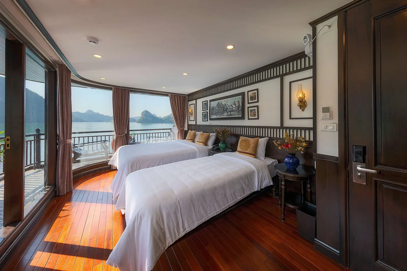 A wood-floored cruise cabin with two white-draped twin beds, dark wood paneling and a curved glass wall opening onto a balcony with a view of calm water and limestone karst islands.