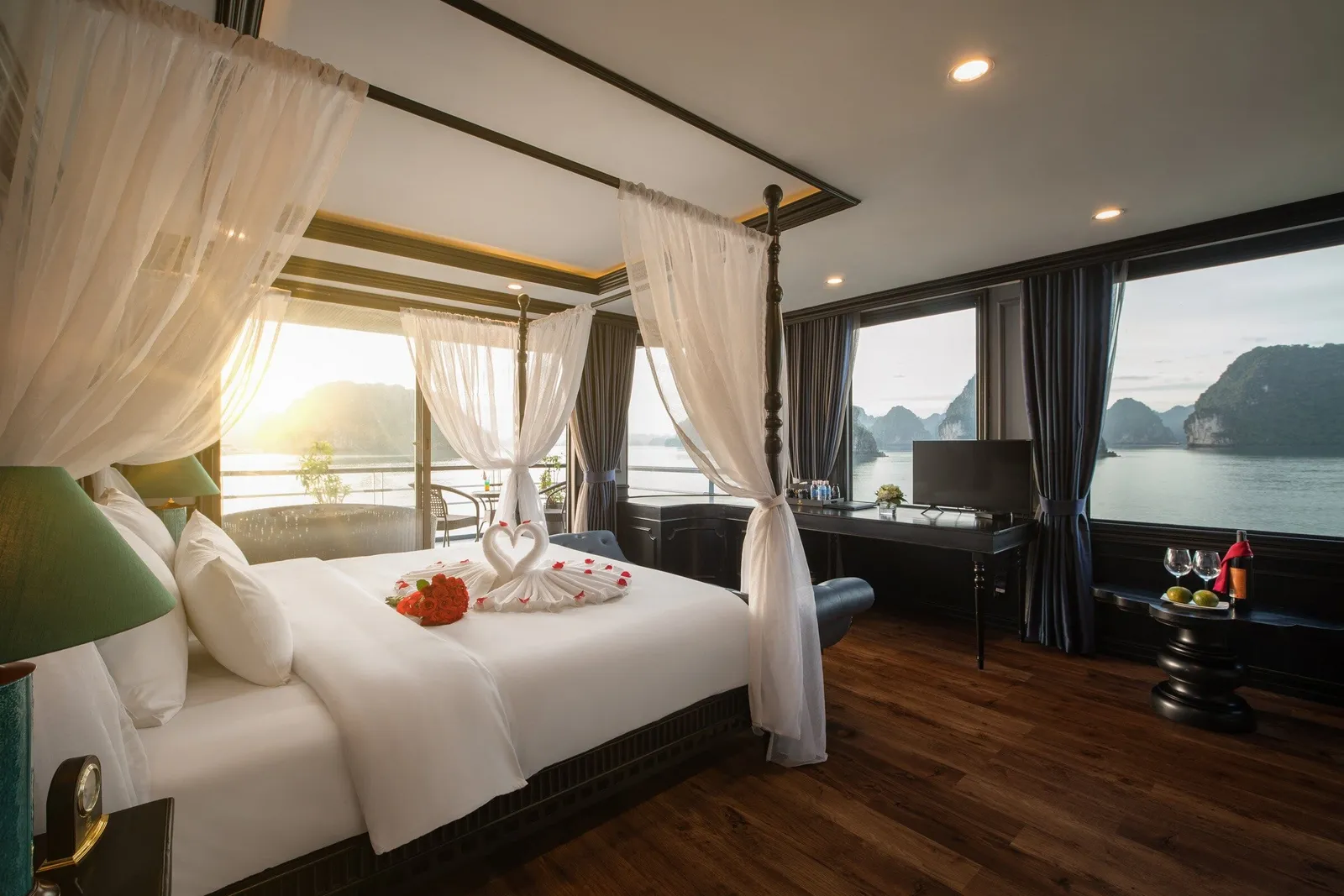 A cruise cabin with a canopy bed decorated with towel swans and red flowers, a desk with TV, and large windows and a balcony overlooking calm water and limestone karst islands at sunset.