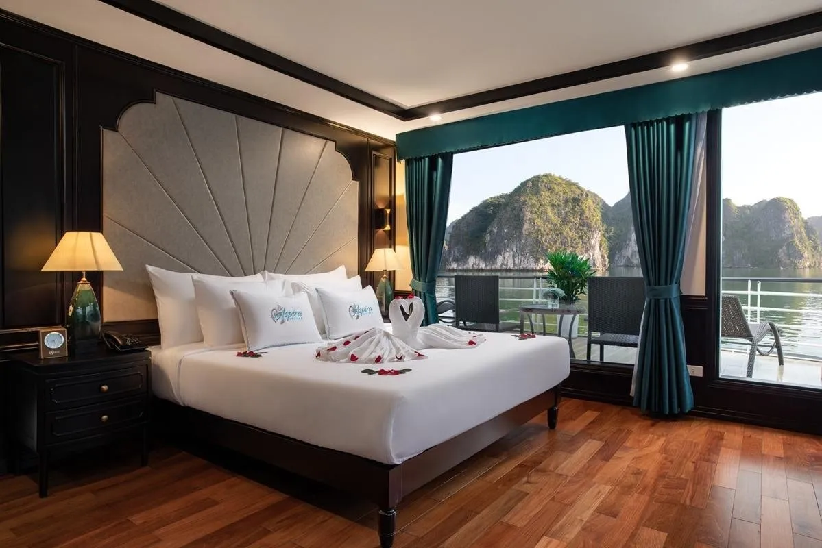 A cruise cabin with a large bed with logo-embroidered pillows and a towel swan with rose petals, dark wood walls, teal curtains, and a balcony window overlooking limestone karsts and calm water.