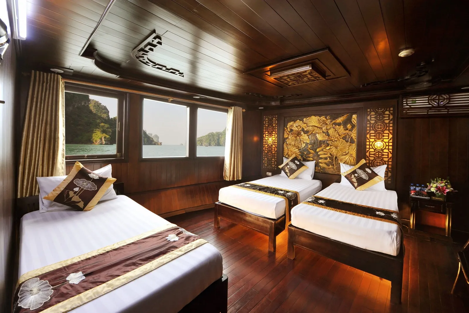 A wood-paneled cruise cabin with three twin beds dressed in white linen and brown-and-gold lotus-pattern runners, with windows showing calm water and green limestone karst islands.