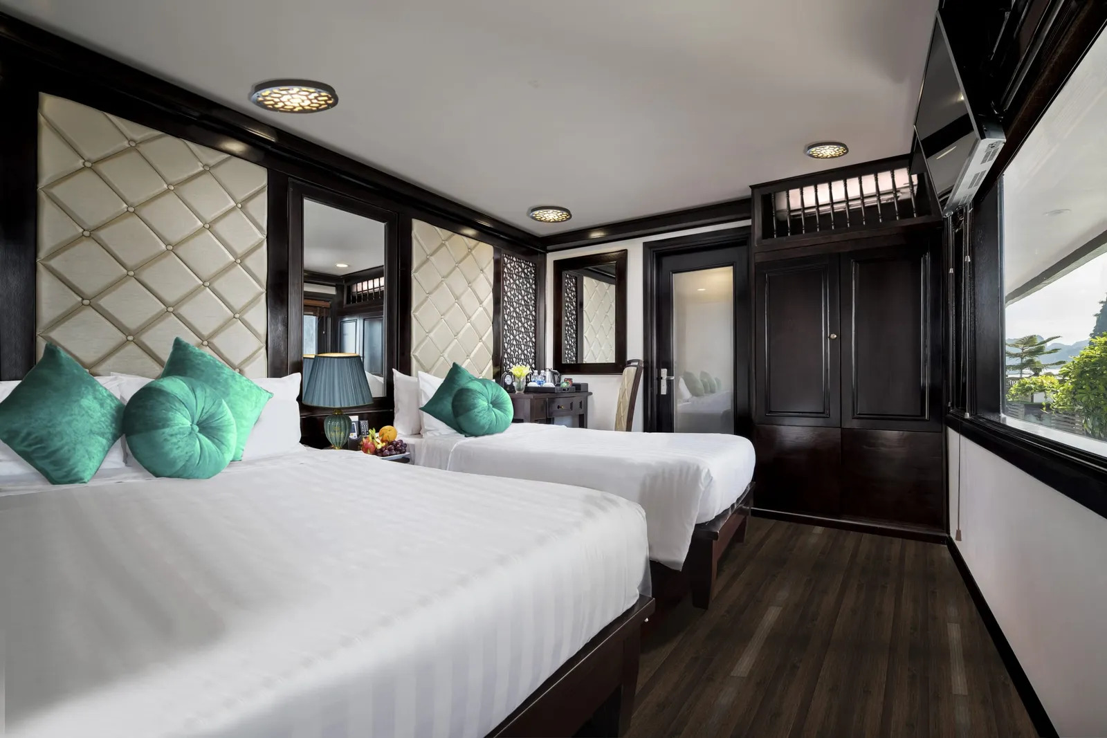 A twin-bed cruise cabin with dark wood panelling, tufted cream headboards, teal cushions, a wall-mounted TV, and a window showing green trees and a distant karst hill.