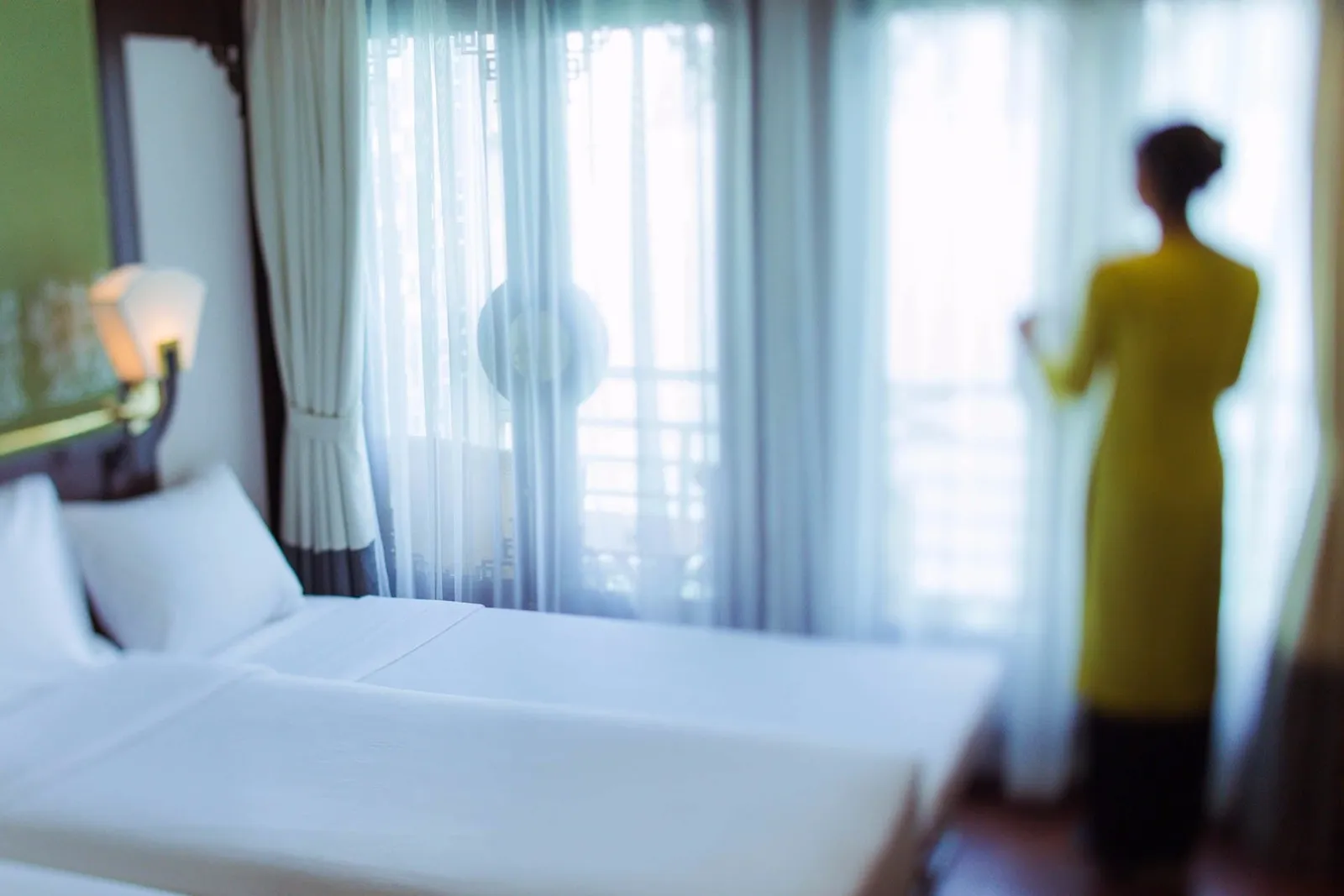 A softly blurred cabin interior with twin white beds, a wall lamp and sheer curtains, while a woman in a yellow ao dai stands at the window with her back to the camera.