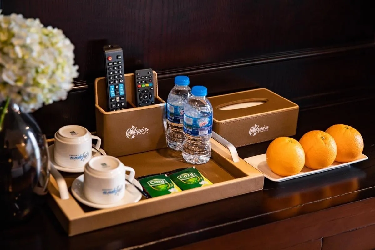 A tan leather tray on a dark wooden cabinet holds two teacups, tea sachets, two water bottles, a remote holder with remotes and a tissue box, both embossed with an Aspira Cruises logo, with a plate of three oranges and white hydrangeas beside it.