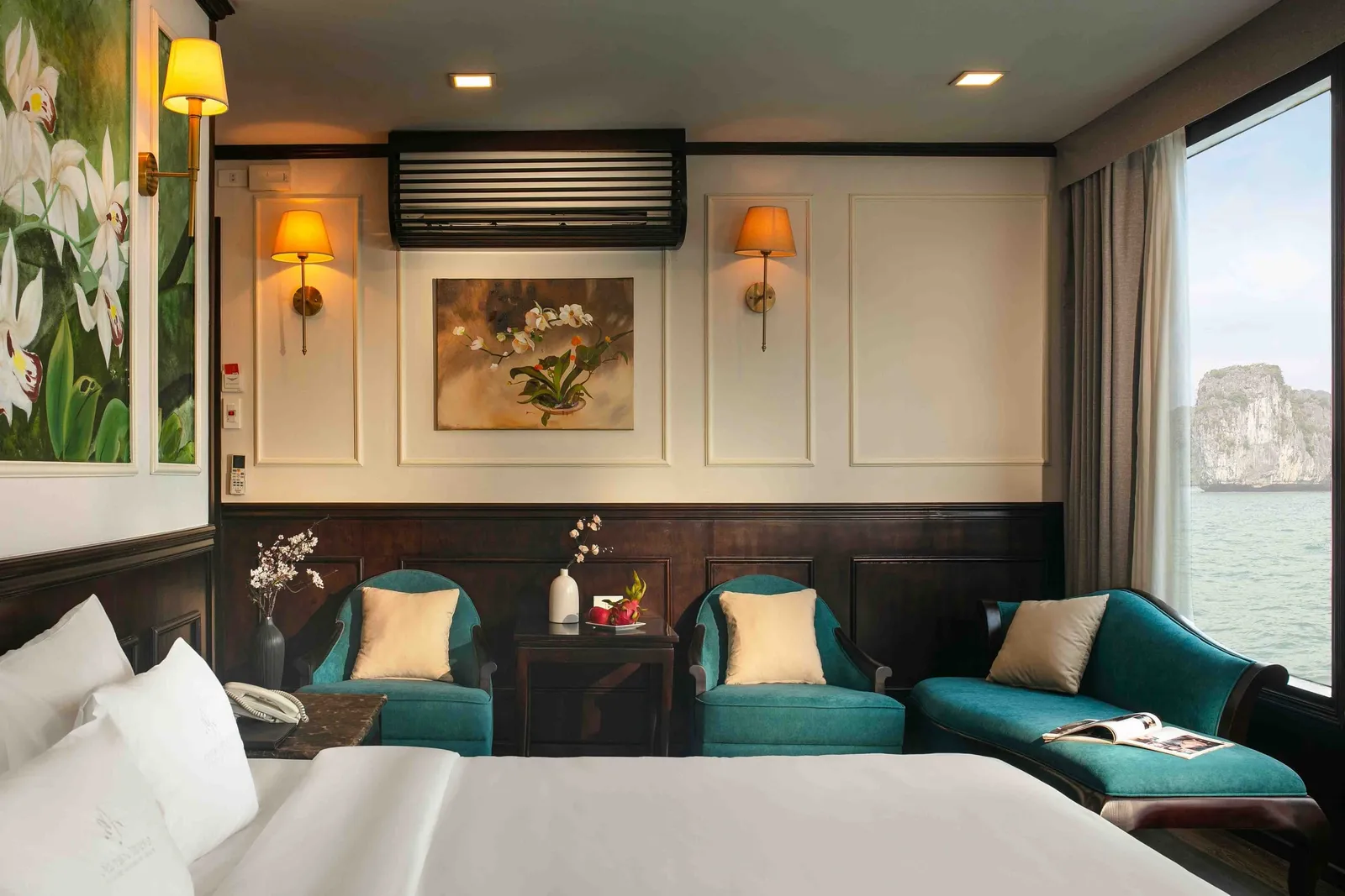 A cruise cabin with a white bed, teal armchairs and chaise, wall sconces and orchid art, with a window showing calm water and a limestone karst island; a faint logo is embroidered on the pillow.