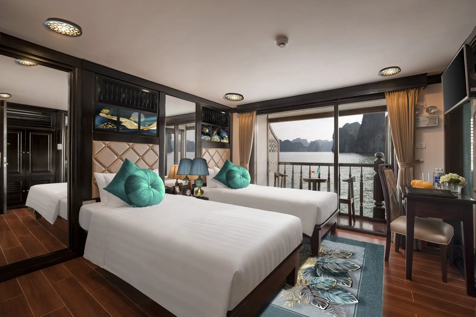 A cruise cabin with two twin beds, teal cushions, dark wood furniture and a mirrored wall, with sliding glass doors opening onto a balcony that overlooks calm water and limestone karst cliffs.