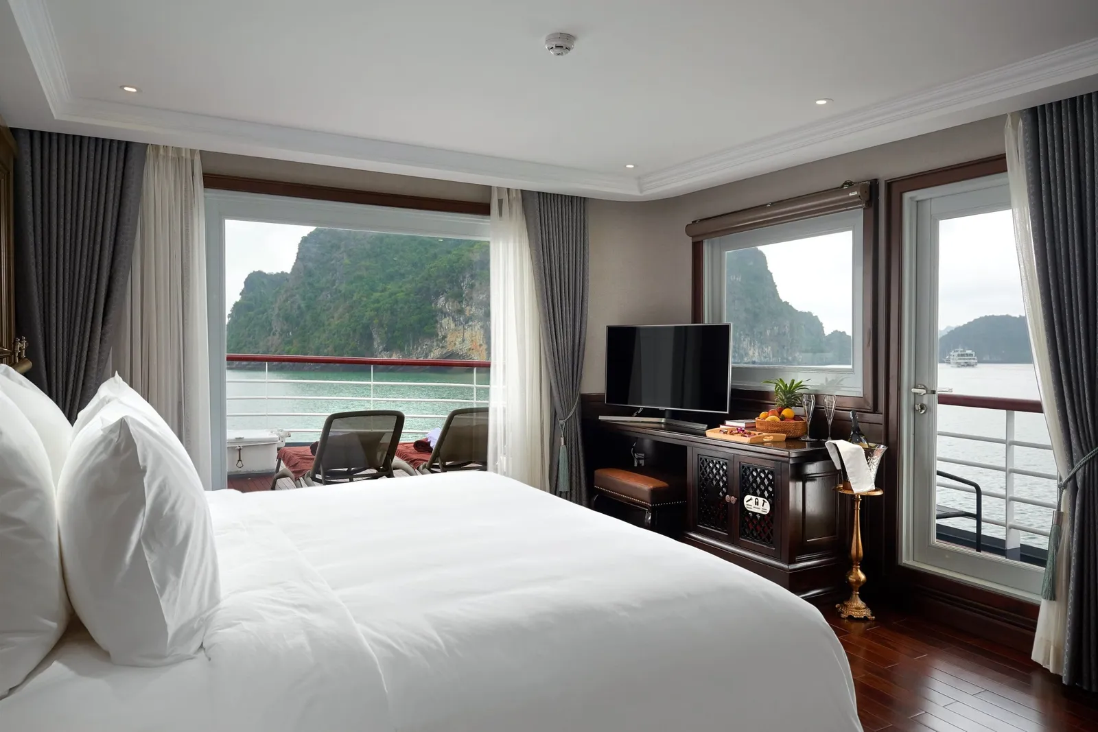 A cruise cabin with a white-bedded double bed, dark wood furniture and a TV, with windows and a glass door opening onto a private balcony with loungers and a view of green limestone karsts over calm water.
