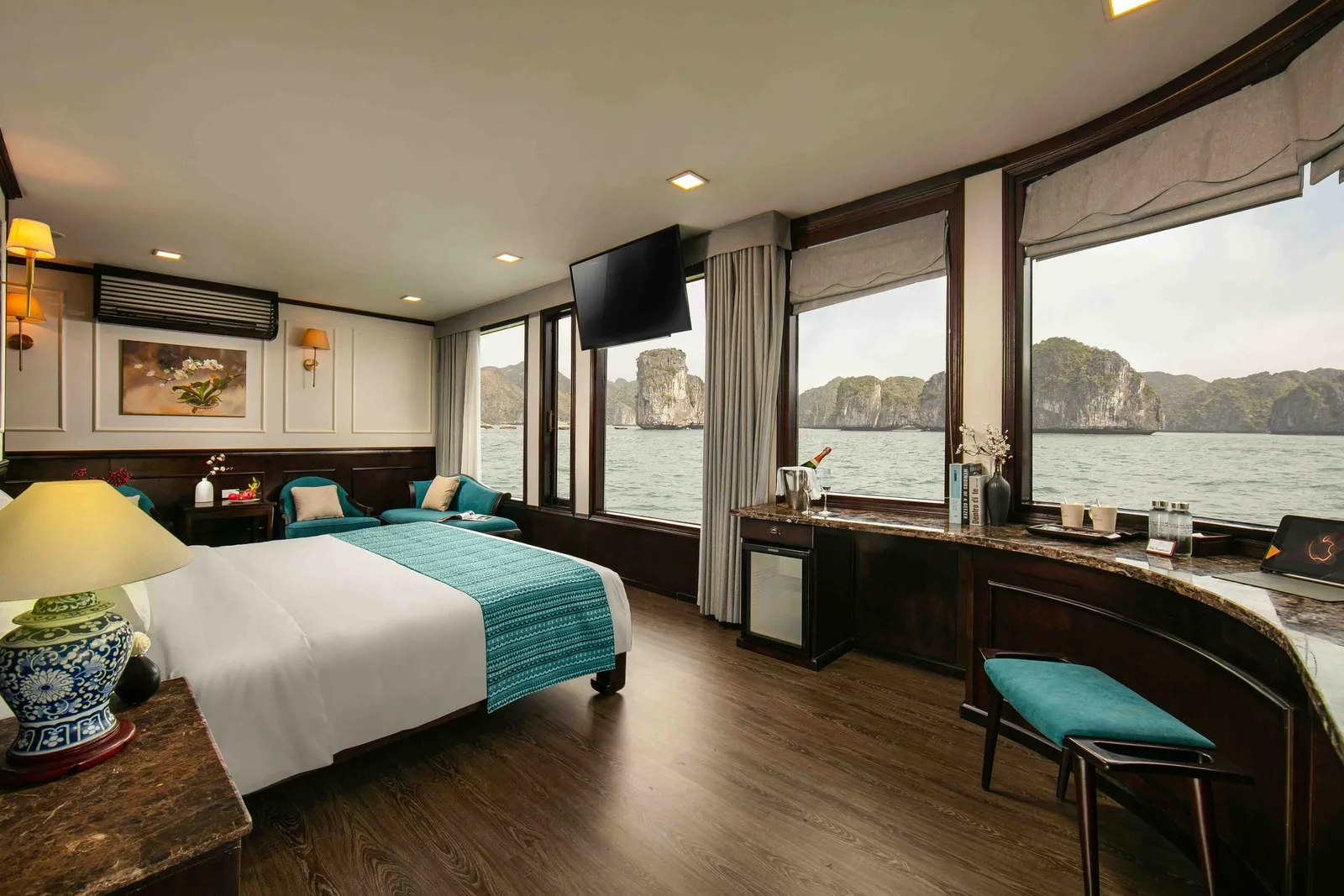 A cruise cabin with a bed, teal accents, a TV and a curved desk, with wide windows showing calm water and limestone karst islands outside.