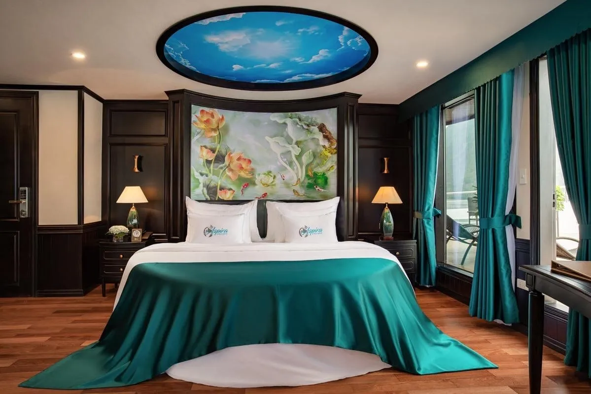 A cruise cabin with a double bed, teal satin runner and curtains, lotus mural headboard, oval sky-image ceiling panel, and pillows embroidered with a logo, with a balcony visible through the glass door.