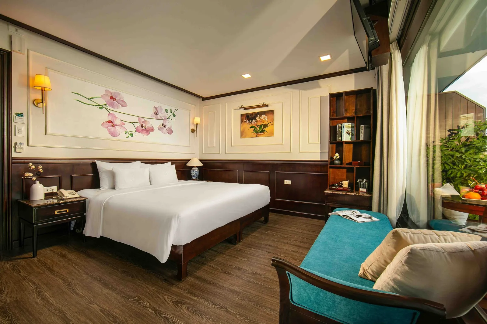 A cabin with a large white-draped bed, dark wood paneling, orchid artwork, a teal sofa, and a large window on the right showing plants and a fruit plate on a balcony table.