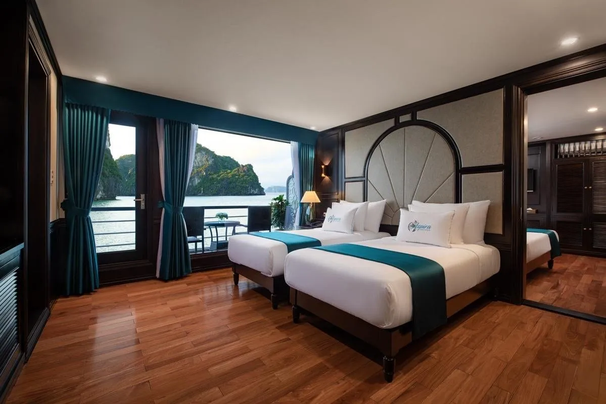 A cabin with two twin beds with teal runners and logo-embroidered pillows, dark wood paneling and teal curtains, with a balcony door opening onto water and limestone karst islands.