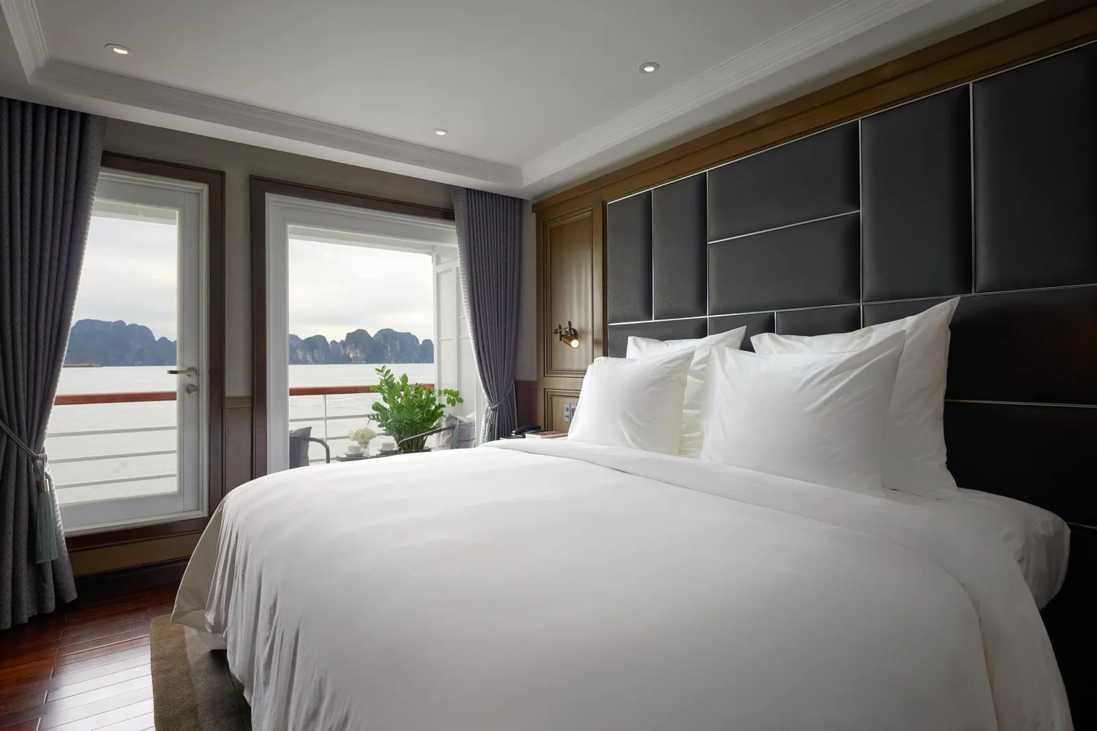 A cabin with a large white bed and dark padded headboard, with glass doors opening onto a balcony that overlooks calm water and limestone karst islands.