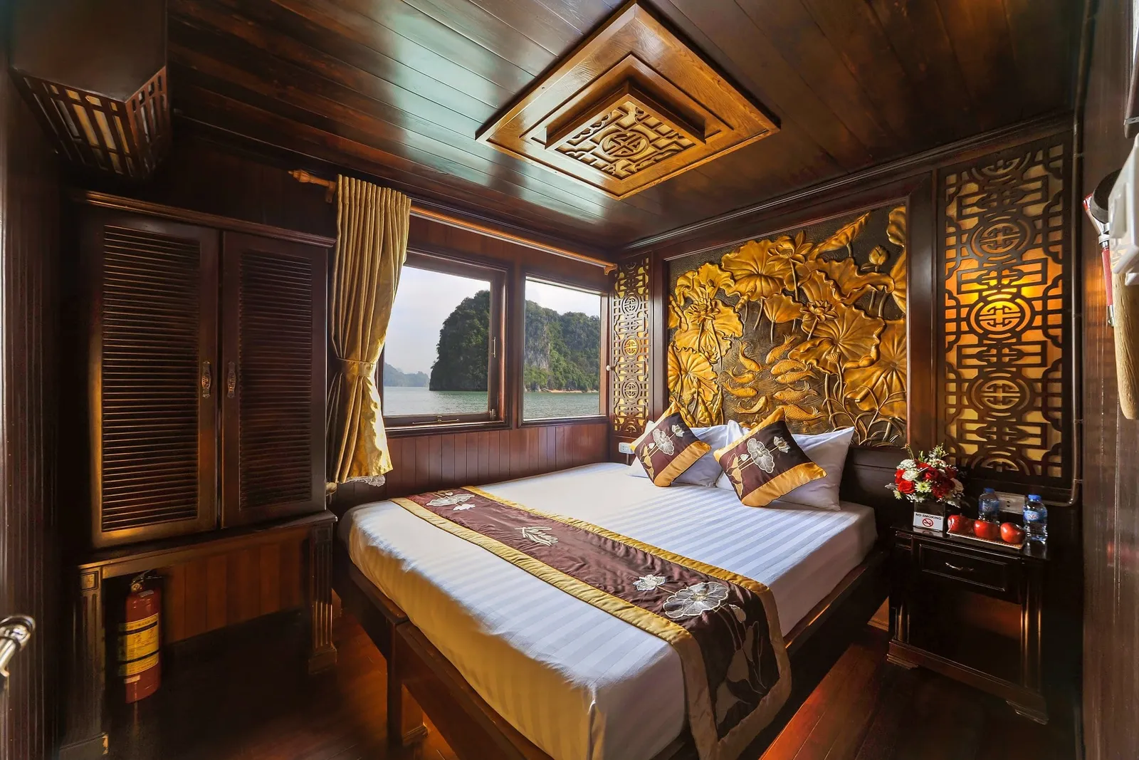 A wood-paneled boat cabin with a double bed, carved golden lotus panel headboard, and a window showing calm water and a limestone karst island.