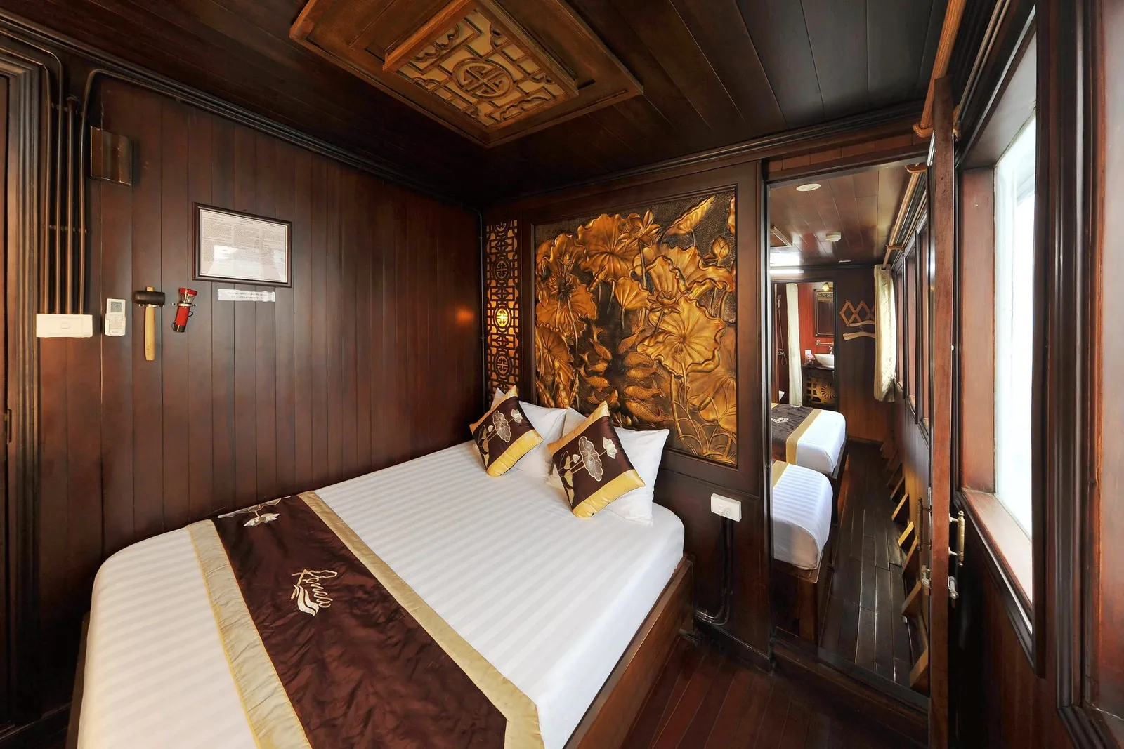 A wood-paneled cabin with a white-sheeted double bed, a brown and gold embroidered runner bearing a script logo, cushions, a carved golden lotus panel, and a mirror reflecting a second bed and the bathroom.