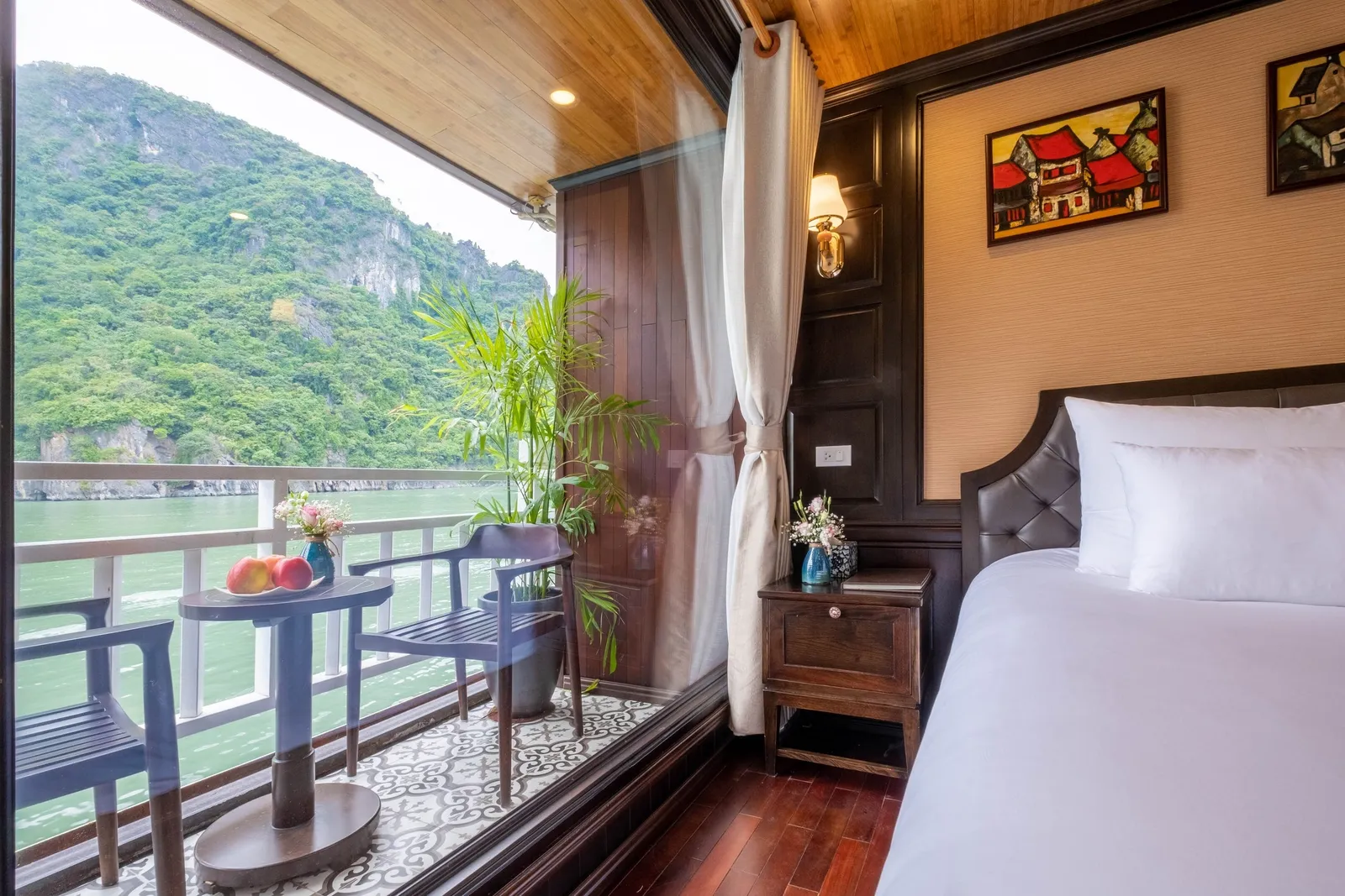 A wood-paneled cruise cabin with a white-draped bed, nightstand and framed paintings, opening through glass onto a small balcony with two chairs, a table with fruit, and a view of green karst cliffs over calm water.
