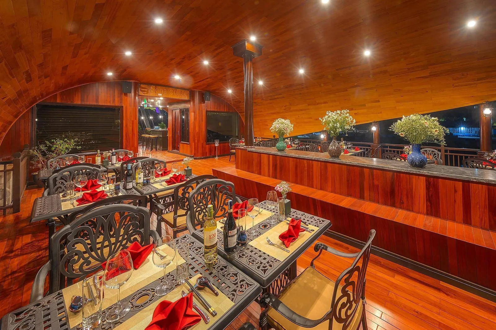 A wood-paneled upper-deck dining area with set tables, red napkins, wine bottles and flower vases under a curved ceiling with recessed lights, and a signboard reading 'Sena Cruises' in the background.