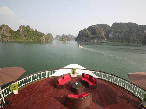 A red-brown wooden upper deck with wicker sofas with red cushions, a round table, brown umbrellas and a white railing, overlooking green water and limestone karst islands with a small boat in the distance.