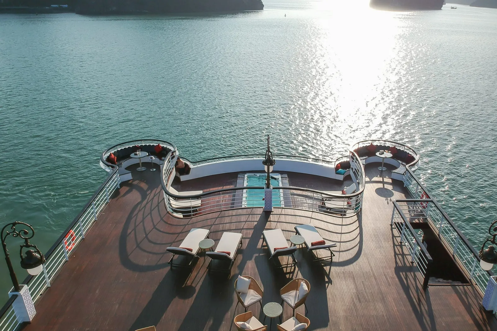 High-angle view of a wooden sun deck with loungers, wicker chairs, curved railings, a small pool and lamp posts, overlooking calm green water with dark karst islands in the distance.