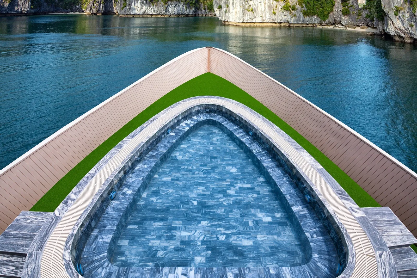 A bow-shaped plunge pool tiled in blue-grey marble sits on the front deck of a boat, surrounded by artificial grass and wood-panelled railings, with calm green-blue water and limestone cliffs beyond.