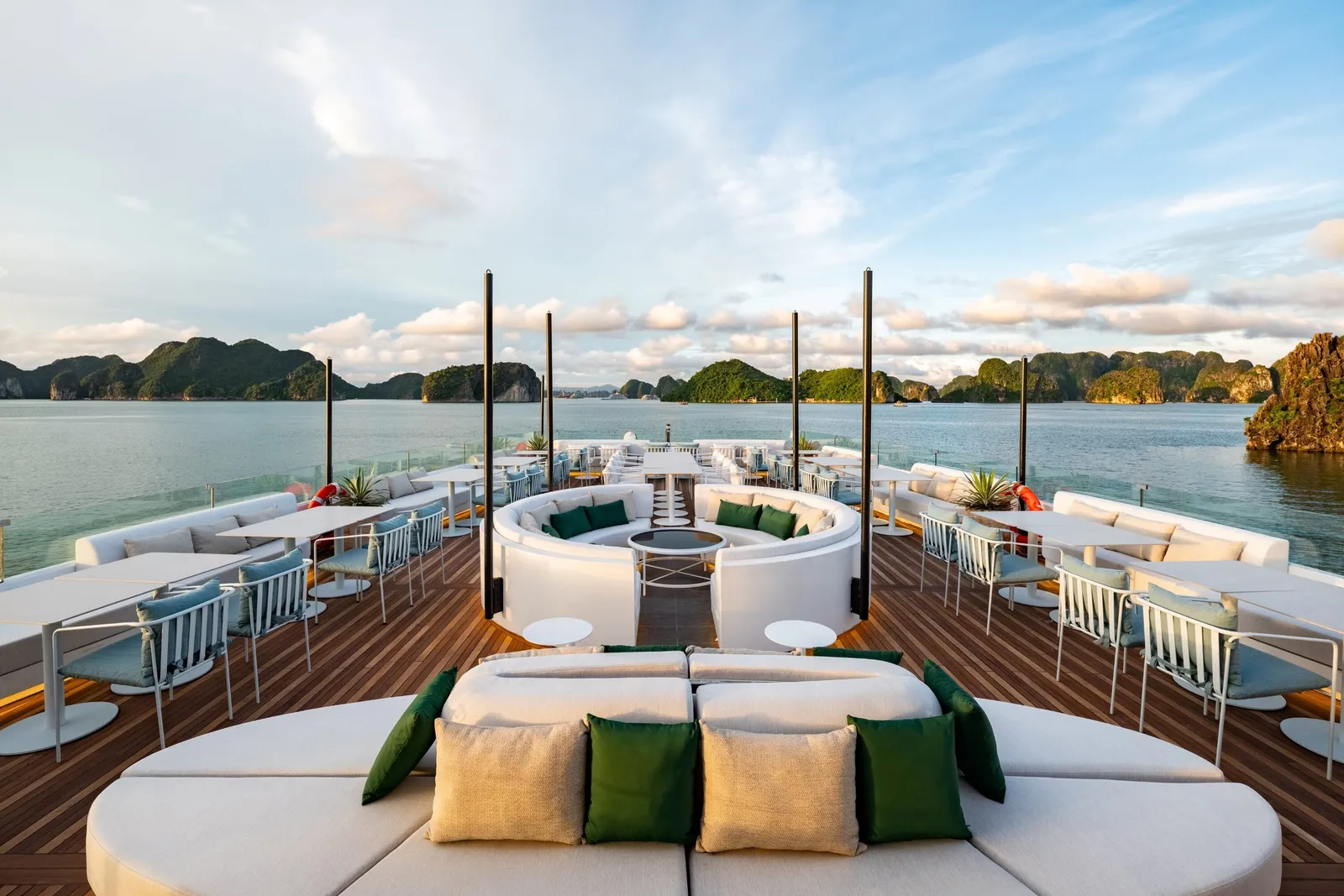 A teak sun deck of a cruise boat with white circular sofas, green and beige cushions, tables and chairs, facing calm water and green limestone karst islands under a blue sky with clouds.