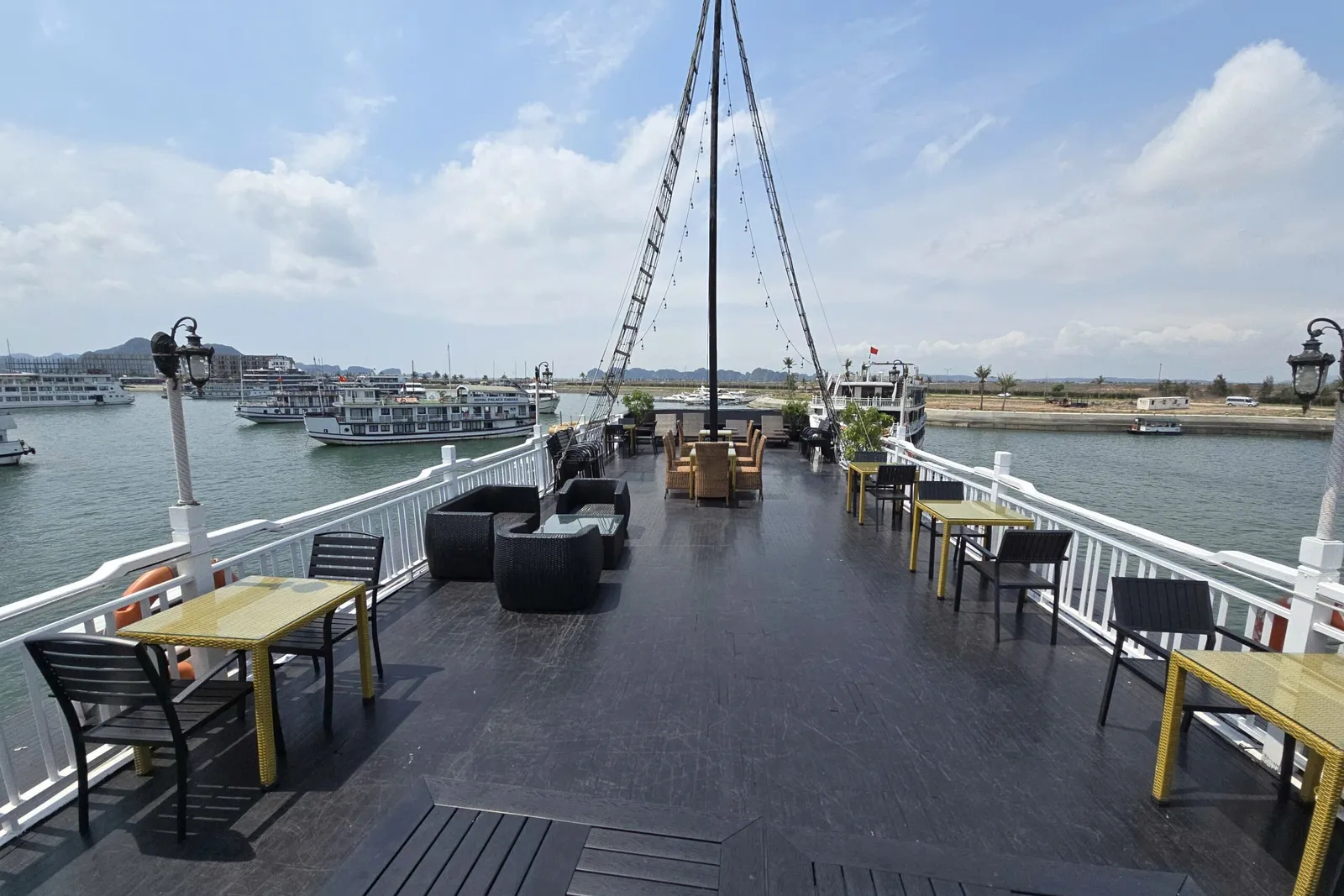 The upper deck of a boat with dark wood flooring, yellow tables, black chairs and lounge seating, white railings and a central mast, looking out over a harbor with other cruise boats and distant karst hills.