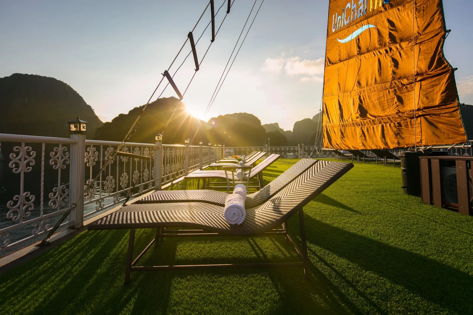 Sun deck with artificial grass, wicker loungers with rolled towels, an ornate white railing and an orange sail printed with 'UniCharm' logo, with limestone karsts silhouetted against a low sun.