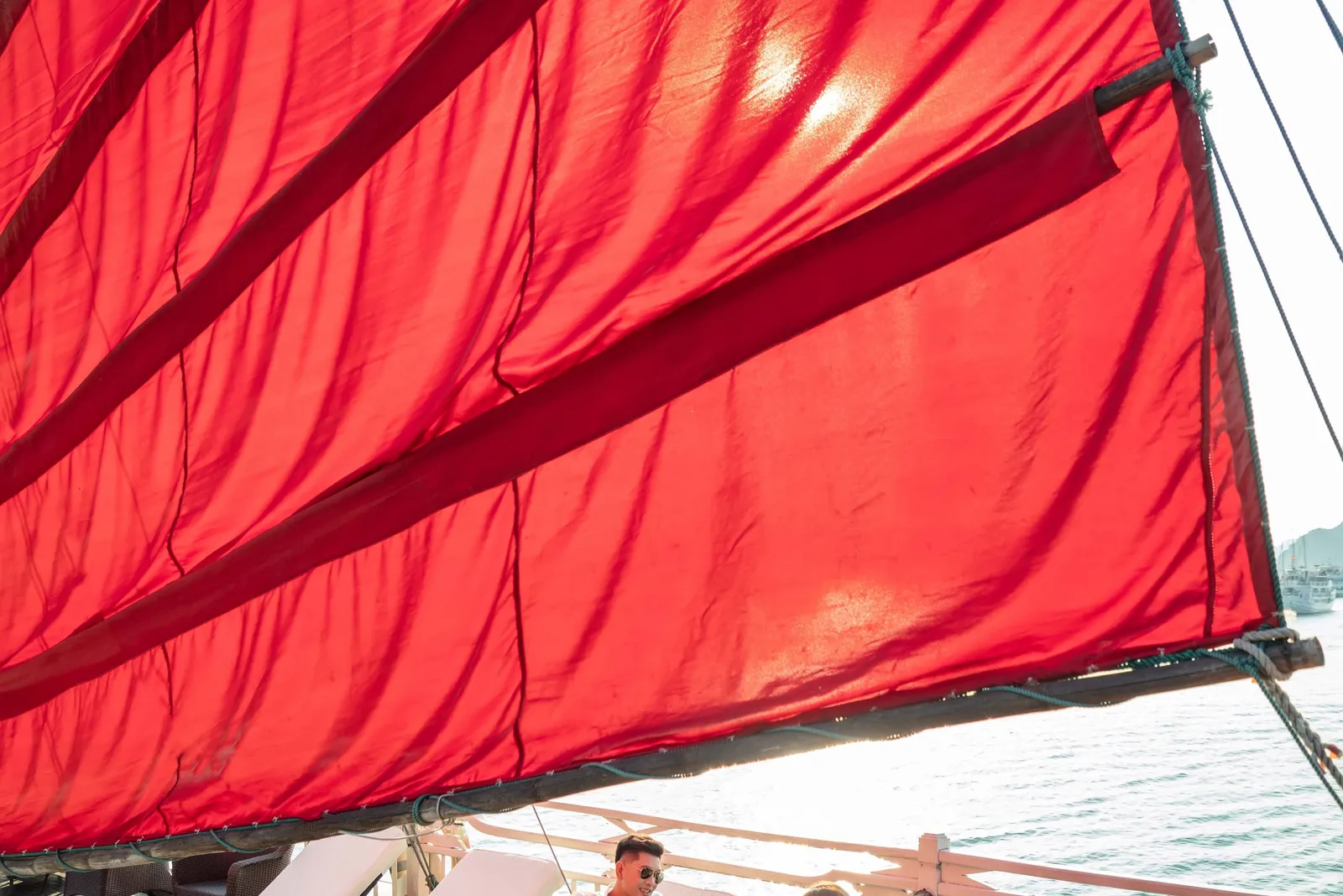 A man and a woman hold wine glasses while sitting on cushioned sun loungers on a dark wooden junk-boat deck beneath a large red sail, with calm water and distant boats and hills behind.