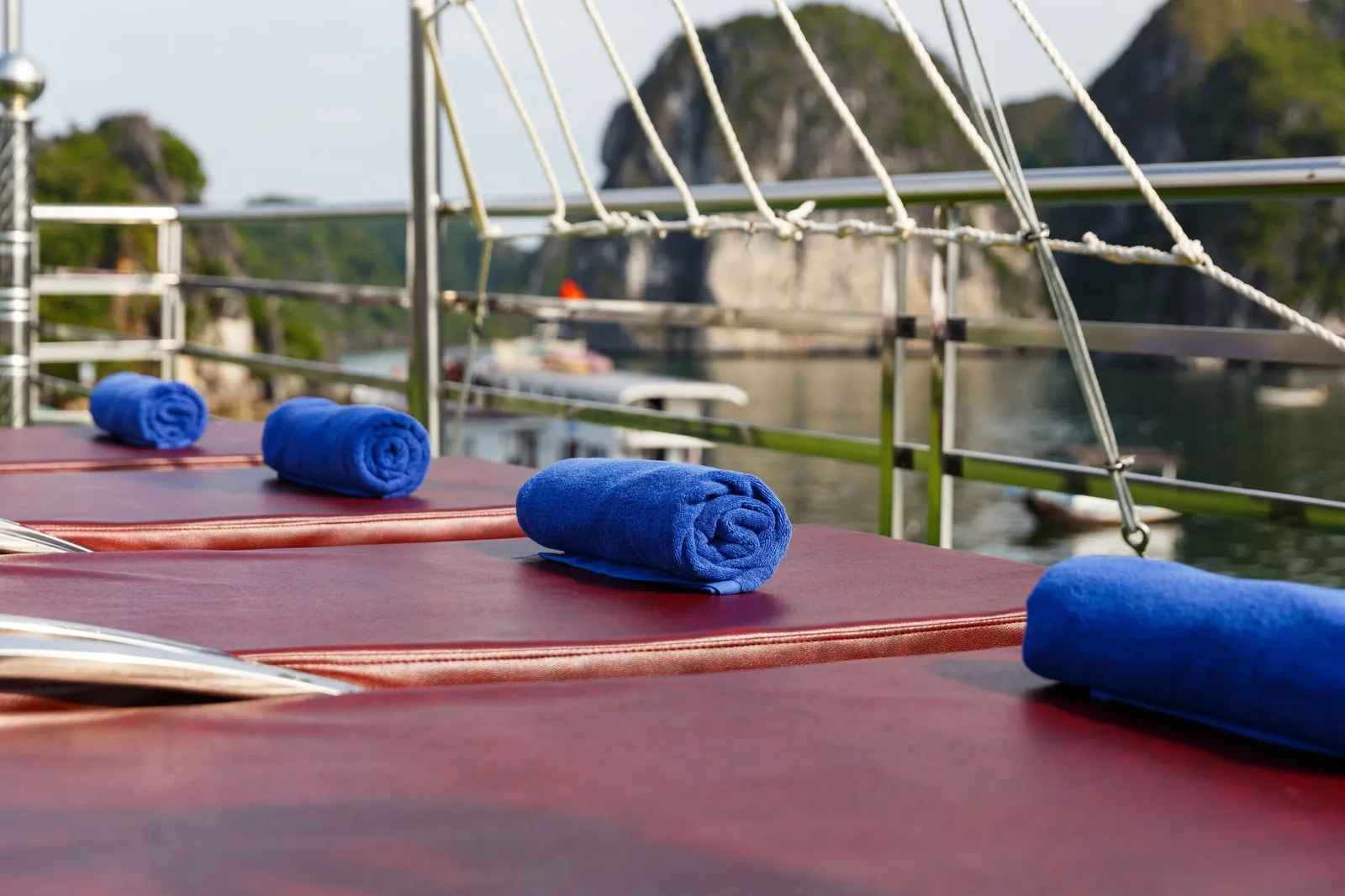 Rolled blue towels sit on red sun loungers on a boat deck with a metal railing and rope netting, with limestone karst cliffs and another boat on the water behind.