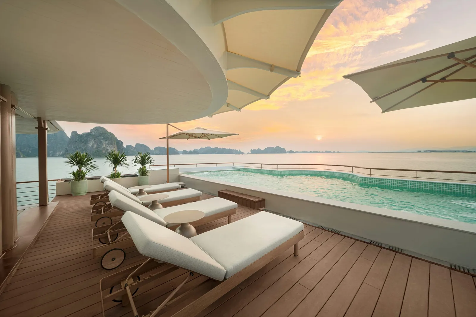 Sun deck of a boat with a row of white sun loungers beside a turquoise pool, shaded by cream canopies and umbrellas, facing calm water and limestone karst islands at sunset.